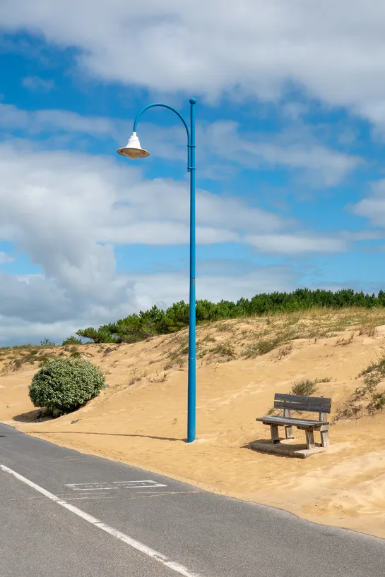 On the side of a small road, a sand dune with a bush, a wooden bench and a blue lamppost