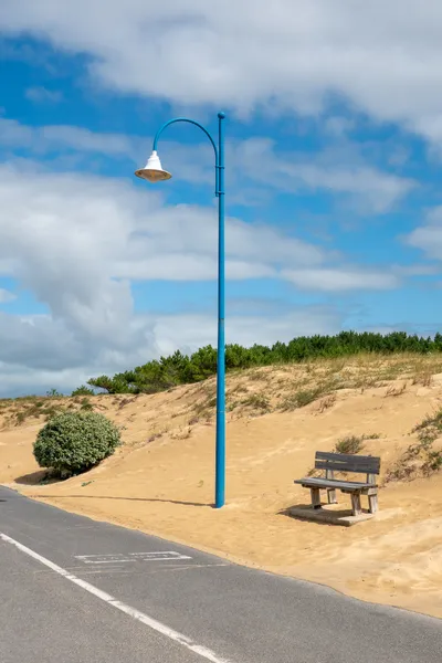 On the side of a small road, a sand dune with a bush, a wooden bench and a blue lamppost