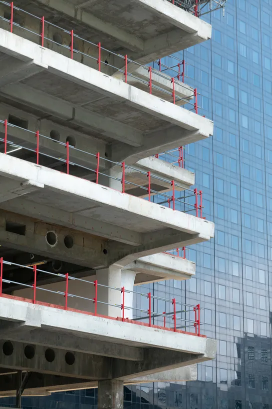 A concrete building being constructed, with bright red fences for falling protection, a modern skyscraper in the background