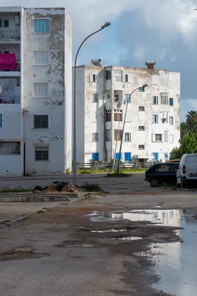 Residential area with tenement buildings, the white paint has been weathered by the years. The road has potholes, in them a puddle reflects the light.