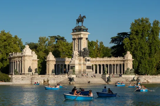 A large stone monument with a bronze statue on top. In the foreground, an artificial lake, with people using rowboats.