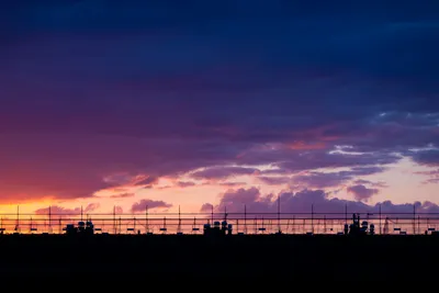 Cloudy sky during sunset, a gradient forms from left to right, bright orange to red to purple. A rooftop in the foreground