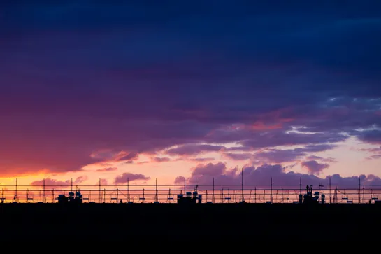 Cloudy sky during sunset, a gradient forms from left to right, bright orange to red to purple. A rooftop in the foreground