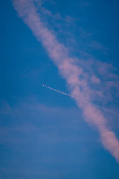 A plane flies through past contrails, both are lit pink by the sunset