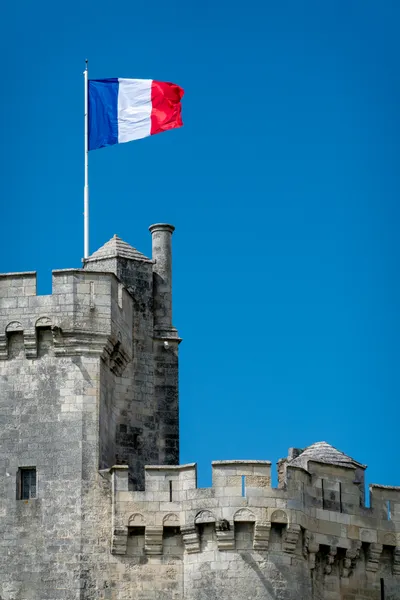 The French flag flies above a stone tower, a clear blue sky behind