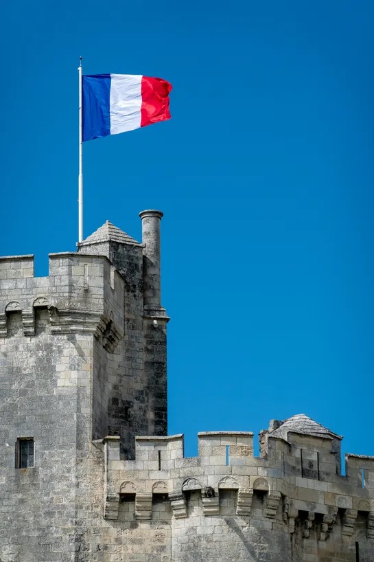 The French flag flies above a stone tower, a clear blue sky behind