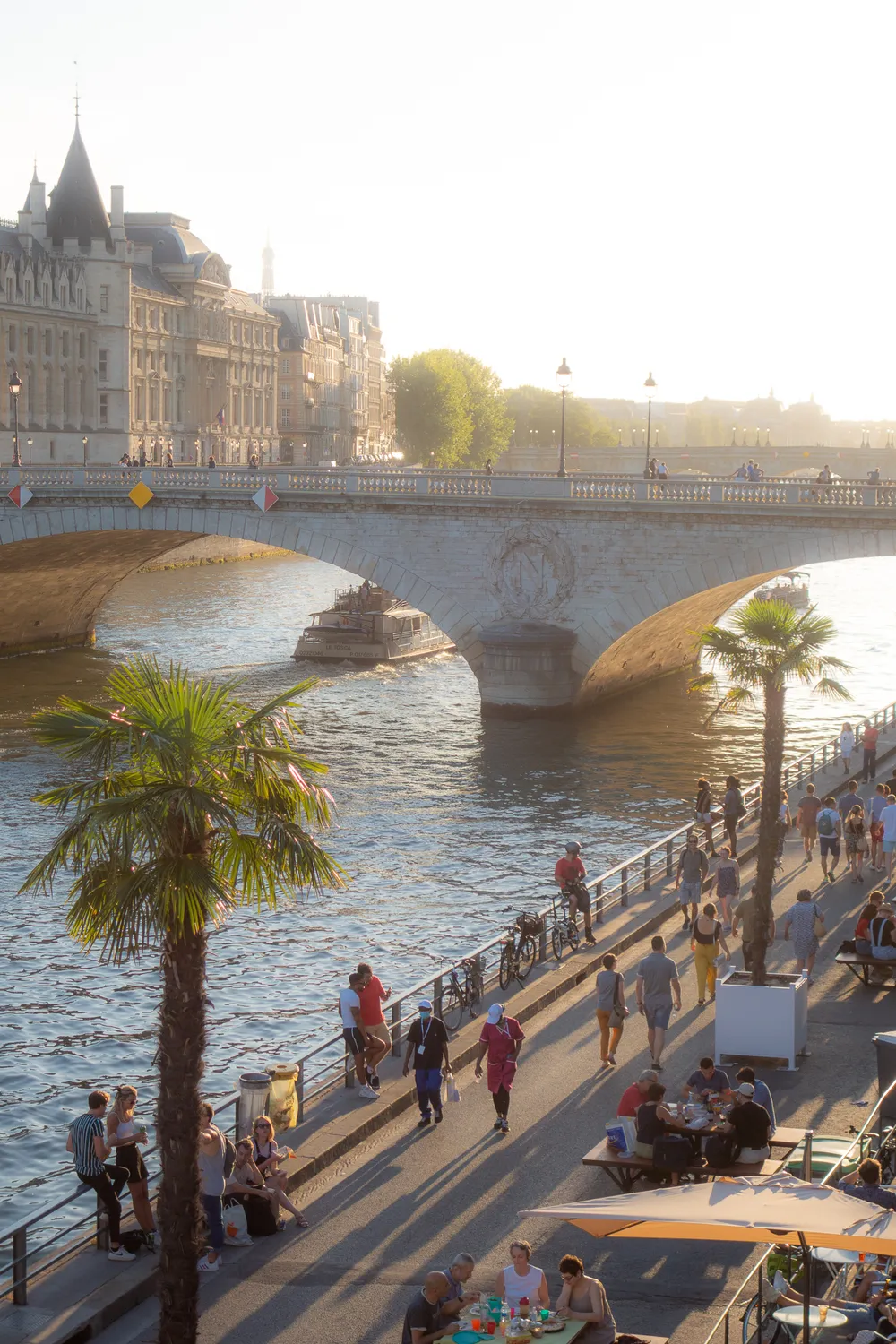 Sunset in the city, on the quays of the river people walking, some sitten down at bars, some palm trees. A stone bridge above the river.
