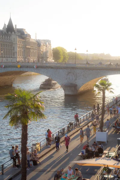 Sunset in the city, on the quays of the river people walking, some sitten down at bars, some palm trees. A stone bridge above the river.