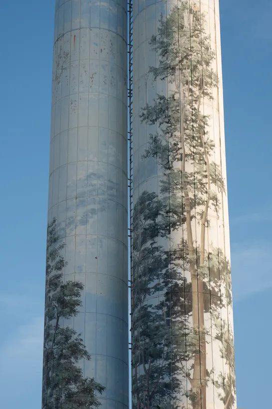 In front of a blue sky, two slim silos, each painted with a blue color and trees, as if to try to hide their presence. The paintings are washed out.
