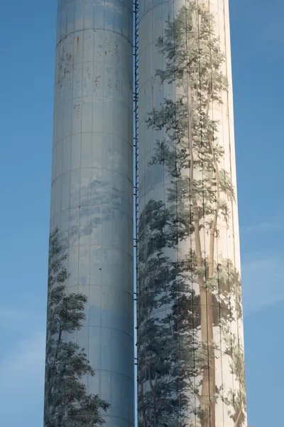 In front of a blue sky, two slim silos, each painted with a blue color and trees, as if to try to hide their presence. The paintings are washed out.