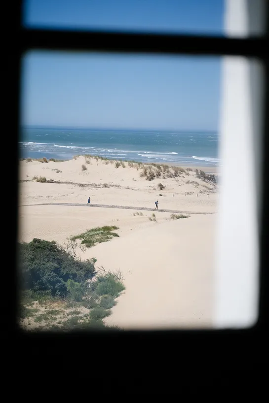 Through a window, a wooden path in the sand with two people waking on it, the sea can be seen further away