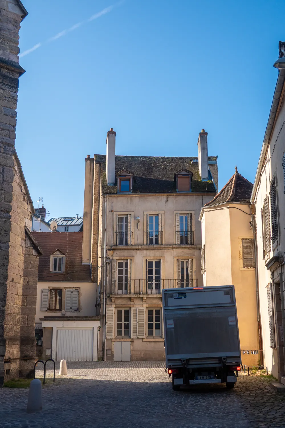 The early morning light hits older buildings in a little street, a great blue sky above, a church to the left, and a moving truck to the right