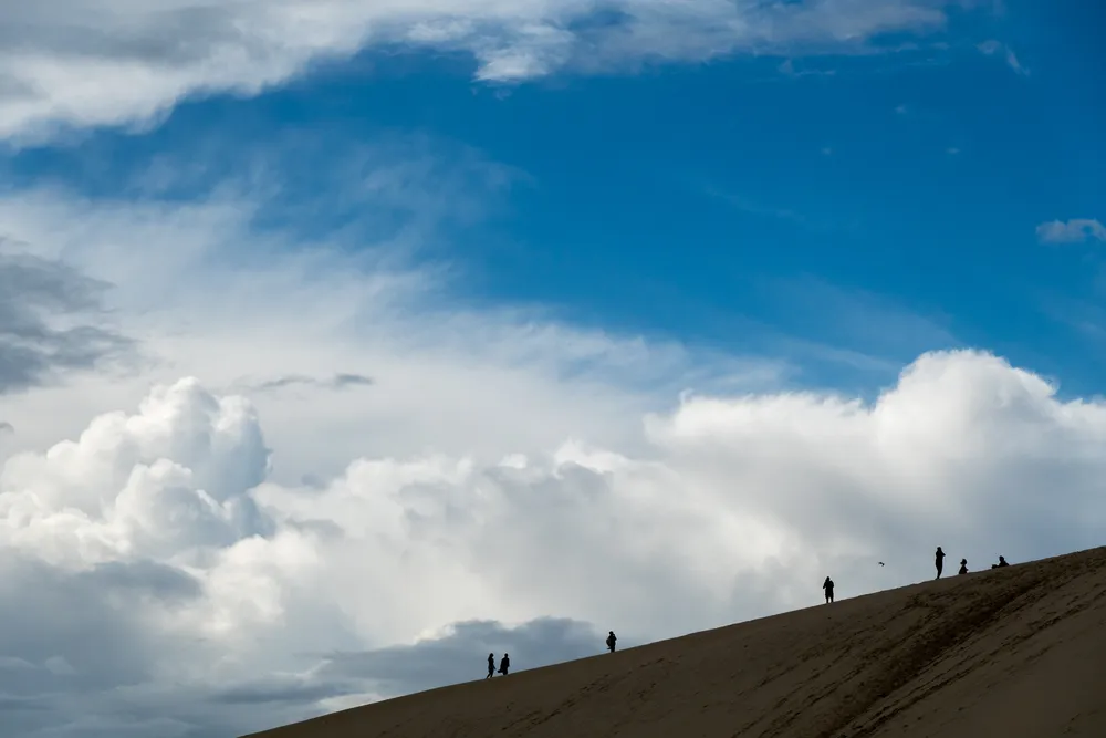 Dramatic clouds in the sky in the background, in the foreground people walking on top of a sandy hill