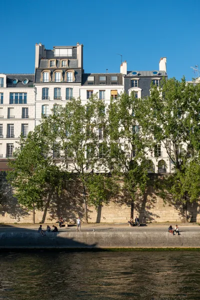 Seen across a river, a quay, people sat on the edge, trees behind, and in the background bright write traditional buildings
