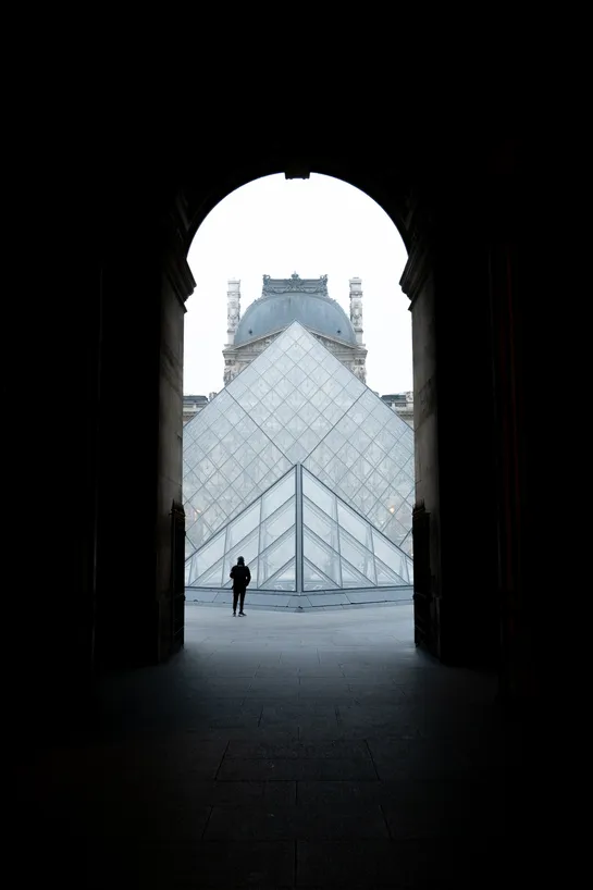 Through a tall tunnel, first a small then the main glass pyramid of the Louvre museum. One person stands in front.