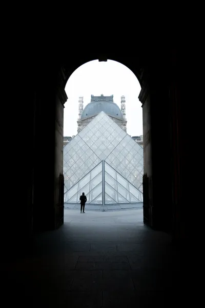 Through a tall tunnel, first a small then the main glass pyramid of the Louvre museum. One person stands in front.