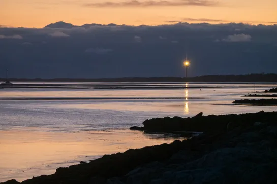 The sea shore, a lighthouse shines on the horizon, cloud cover behind
