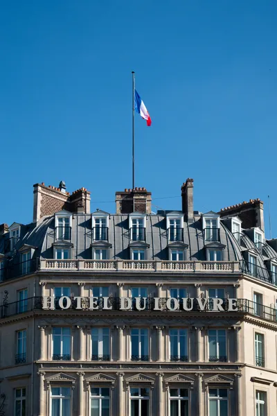 The front of a haussmanian building, a large signage spells "Hotel du Louvre" as a French flag floats above held by a mast