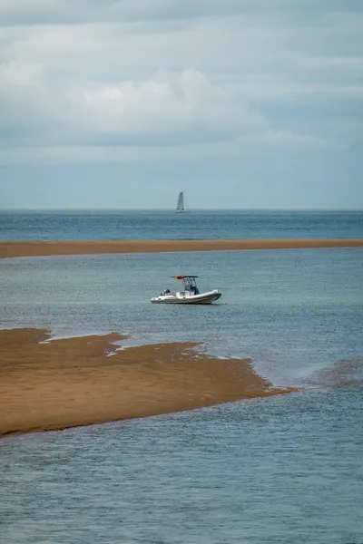 A small boat anchored in a small bay formed by a sand bank, a light house on the horizon