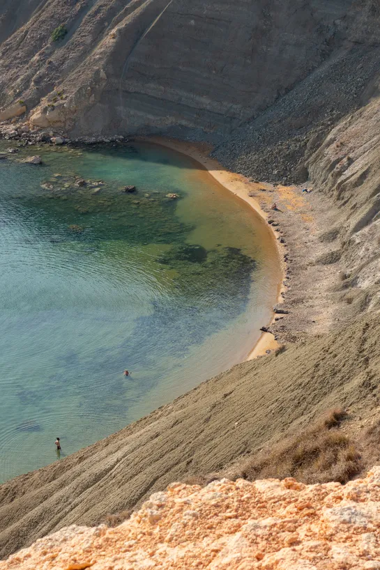 A small bay at the bottom of a cliff, some tourists bathing below in the turquoise waters