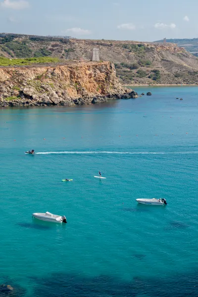 The coastline, a stone tower sits atop a cliff, a jetski comes across the picture. Below are two people paddling in the turquoise waters, near anchored motorboats.