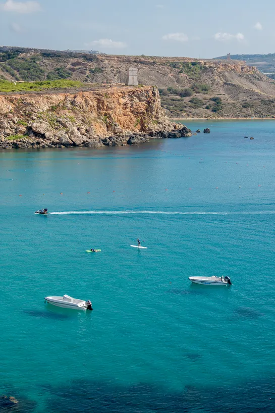 The coastline, a stone tower sits atop a cliff, a jetski comes across the picture. Below are two people paddling in the turquoise waters, near anchored motorboats.