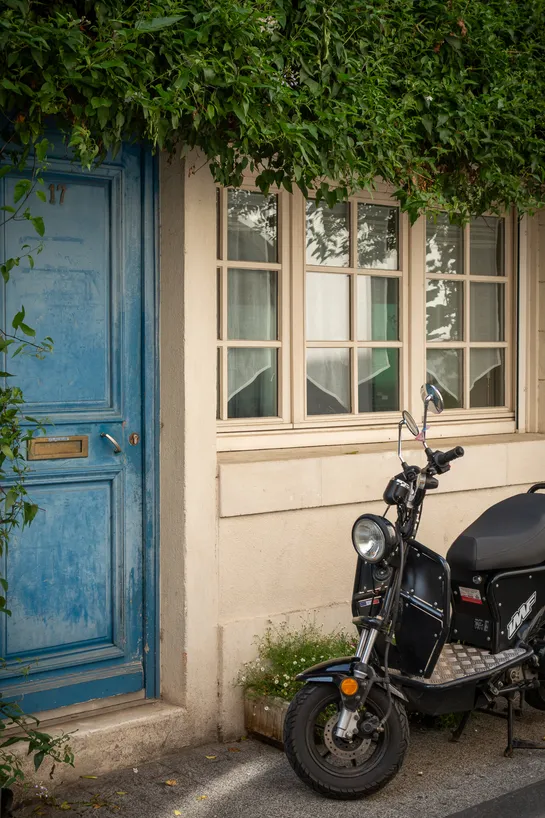 A motorcycle parked in front of a house with a blue door, small windows, and overgrown floiage above