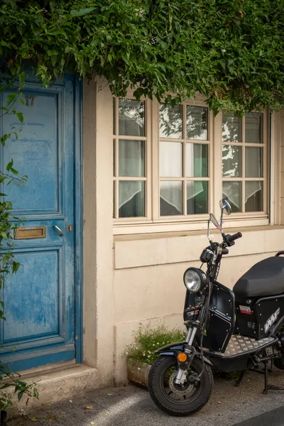 A motorcycle parked in front of a house with a blue door, small windows, and overgrown floiage above