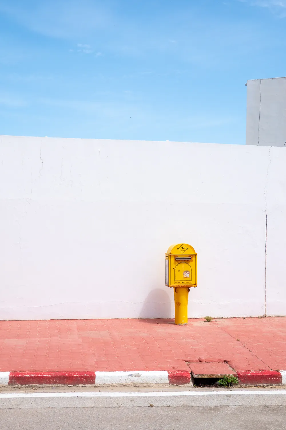 Bright sunlight bathes the scene: a yellow mailbox in front of a plain white wall, atop a red pavement and beneath a pale blue sky.