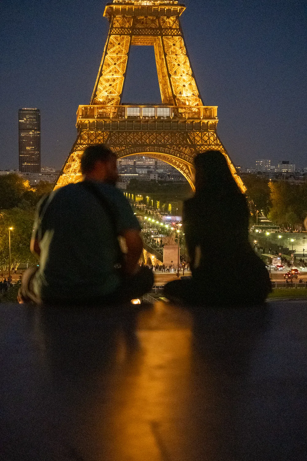 A couple sitting on steps at night, overlooking the Eiffel Tower, the Champ de Mars, and farther away, the Montparnasse Tower