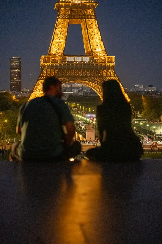 A couple sitting on steps at night, overlooking the Eiffel Tower, the Champ de Mars, and farther away, the Montparnasse Tower