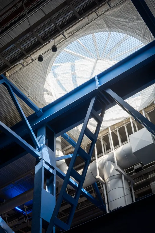 The ceiling of an industrial looking building, with blue metal beams lit by a round opening, the sky can be seen through it
