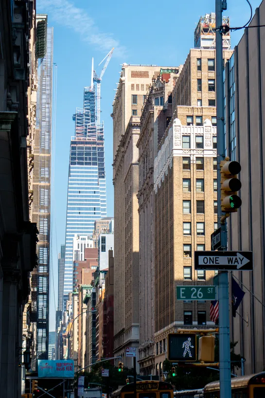 The last floors and spire of a skyscraper being built in the background, a street with other tall buildings on either side of it, and in the foreground is classic New York City signage