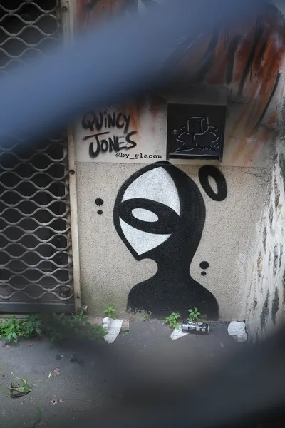 A graffiti of a cartoon figure, with a sideways eye as a head, on a wall in a neglected part of a street