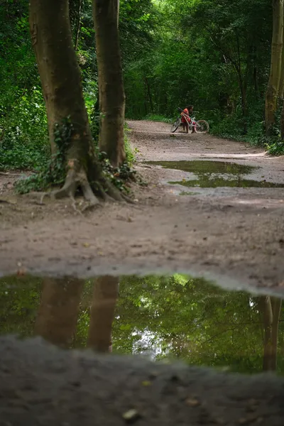 The pathway in front of us has large puddles reflecting the forest around us, with some light coming through. In the distance, a man dressed in orange sits with his bike nearby