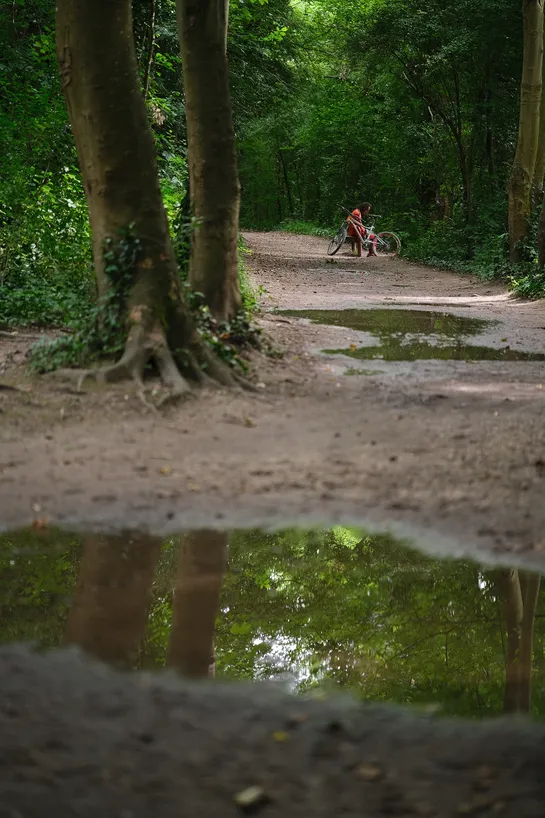 The pathway in front of us has large puddles reflecting the forest around us, with some light coming through. In the distance, a man dressed in orange sits with his bike nearby