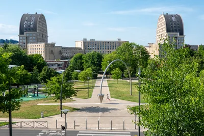 Two identical rounded buildings sit across each other, with a large plaza and trees between them, a walkway coming to us, with a child playing