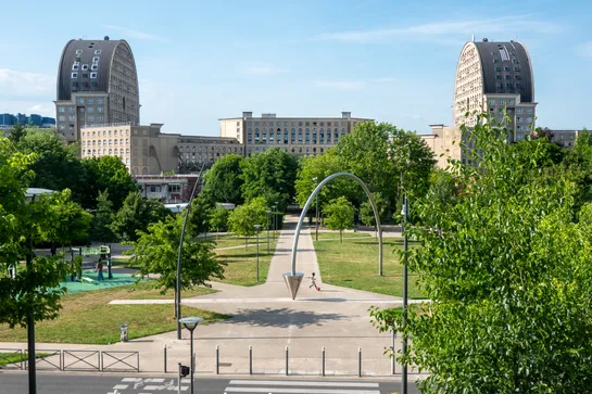Two identical rounded buildings sit across each other, with a large plaza and trees between them, a walkway coming to us, with a child playing