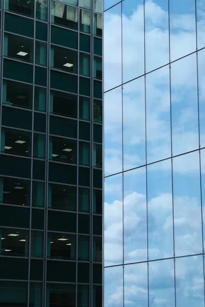 The frame is split in two along the vertical axis, on each side a skyscraper from up close, on the left, office rooms with standard large lighting peaking through the windows, on the right, only the clouds and sky are visible in the reflection