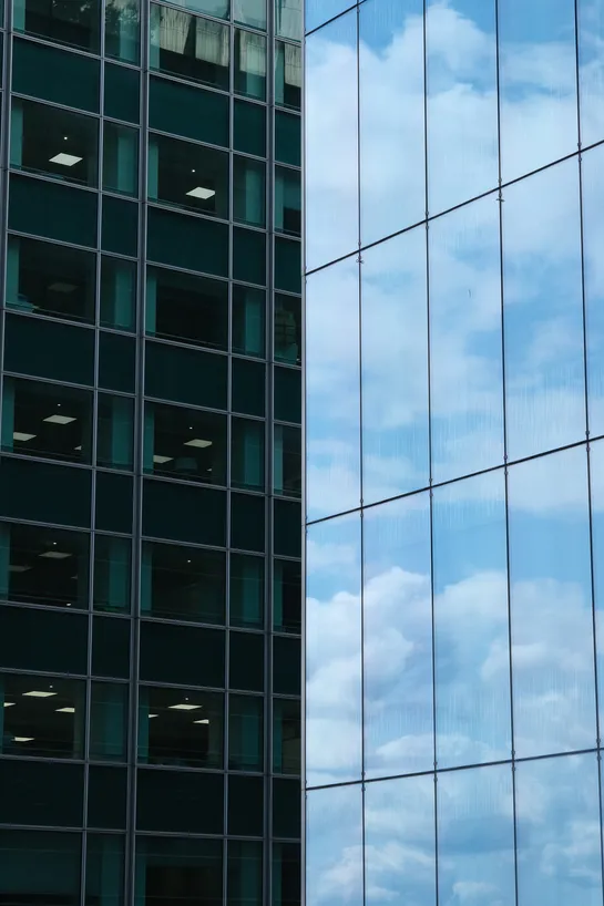 The frame is split in two along the vertical axis, on each side a skyscraper from up close, on the left, office rooms with standard large lighting peaking through the windows, on the right, only the clouds and sky are visible in the reflection