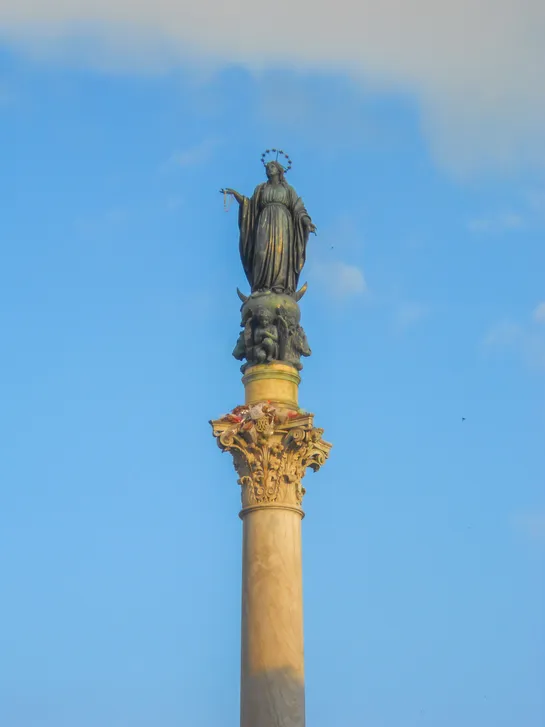 A green statue sits atop a marble column in front of a light blue sky, lit by sunset