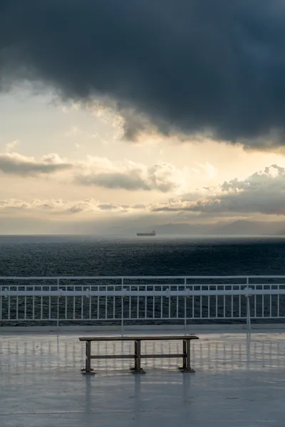 A lone bench before white railings, in the background the open sea, dramatic light and clouds and a single large ship looking small on the horizon