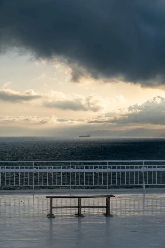 A lone bench before white railings, in the background the open sea, dramatic light and clouds and a single large ship looking small on the horizon
