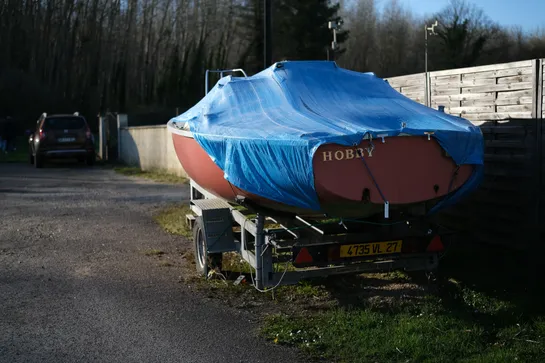 A small boat on a trailer beside a small coutnry road, covered by a blue tarp, the name "Hobby" appears at the back of the boat