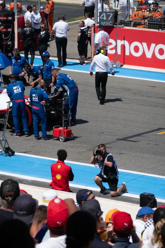 The final moments of preparation before a Formula 1 race, spectators looking on mechanics preparing cars, an FIA official looking back. In the center, Mattia Binotto stands in his bright red Ferrari clothes, being photographed by a journalist.