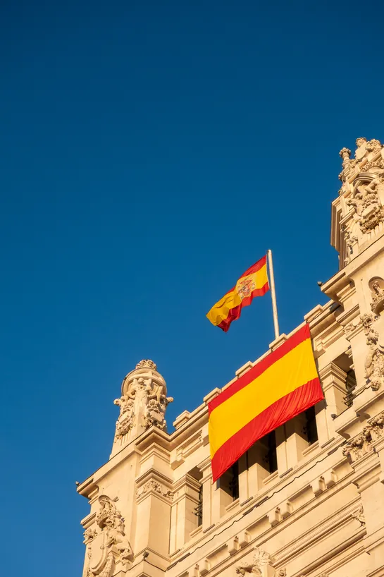 A deep blue sky above, a large stone monument. Two Spanish flags are displayed, one hangs on the monument itself, the other is flies on a mast.