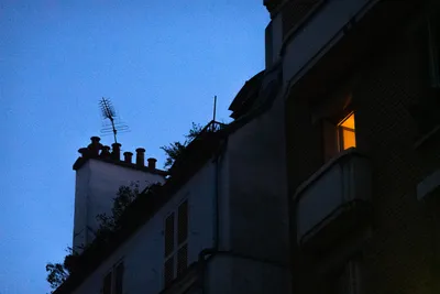 During dusk, rooftops with a single window lit by a yellow lamp