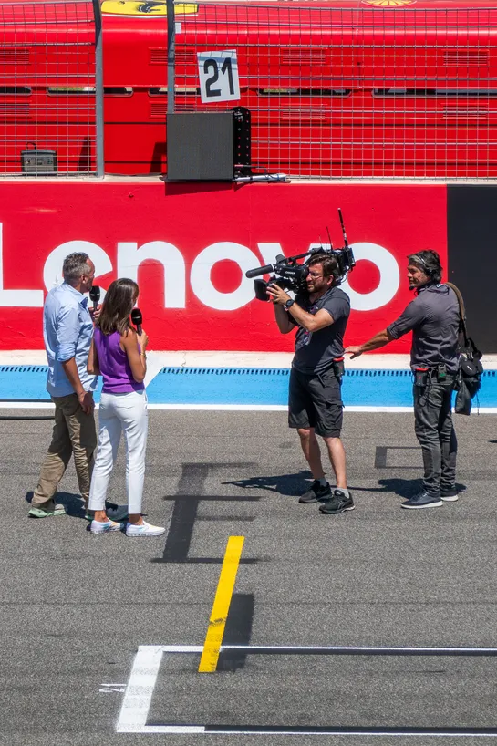 Standing on a race track, two journalists are speaking in front of a cameraman. He is being lightly pulled by a coworker.