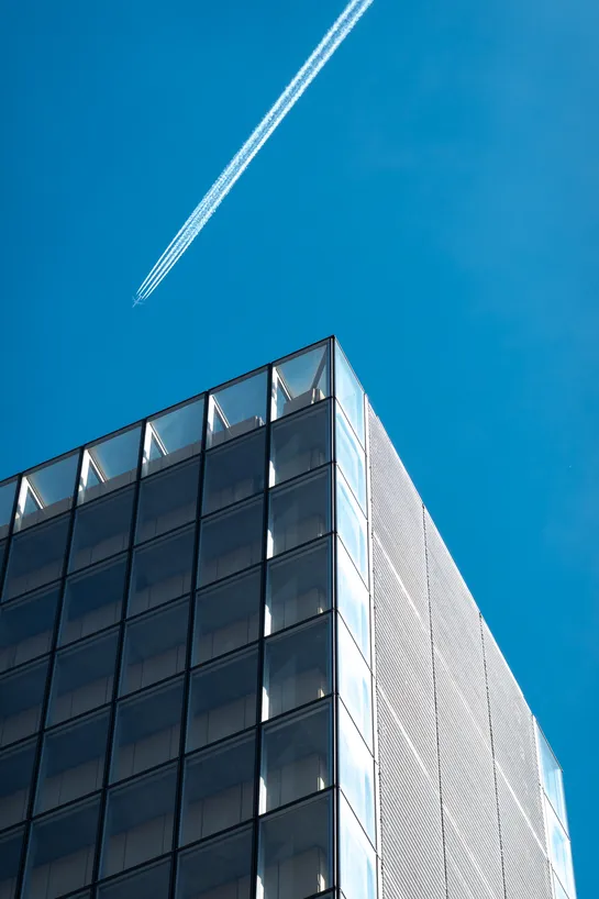 A modern building brightly lit on its side by the sun. In the sky above, a plane leave a very large trail.