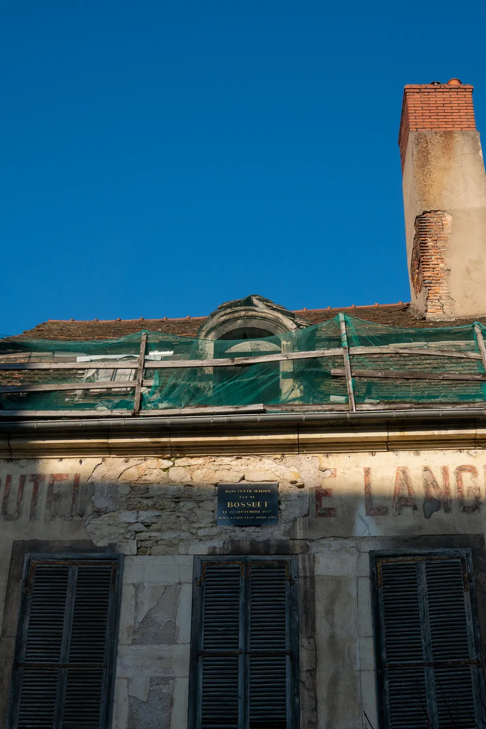 A derelict building roof and chimney, with a flimsy net to prevent the fall of debris. The building front has old painted text and a plaque at the center about its past inhabitant.